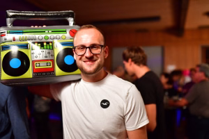 Lächelnder Mann hält eine leuchtend grüne Party-Box vor sich; bunte Bedienelemente und Lautsprecher-Optik, im Hintergrund eine Feiergruppe.