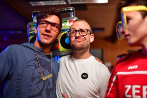 Zwei Männer mit Brillen posieren in einem Raum, im Hintergrund bunte Automaten; Schulter an Schulter, Blick in die Kamera.