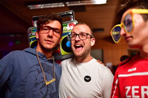 Zwei Männer in Brillen, lachend und nebeneinander; bunte Spielautomatengrafik im Hintergrund, dekoratives Festfoto mit Party-Stimmung.
