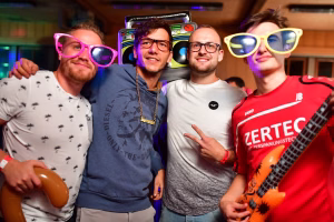 Vier Personen posieren eng beieinander in Partybeleuchtung; alle tragen auffällige Brillen, halten Luftballons und lächeln.