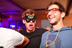 Zwei Männer bei einer Feier, einer mit Bat-Man-Maske, beide lachend; Partylicht im Hintergrund deutet Stimmung und Spaß an.