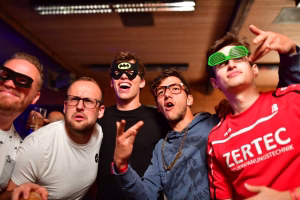 Fünf Personen posieren in einem Raum mit Party-Stimmung: Maskenbrillen und eine bunte Augenmaske, ein Glas in der Hand, lachende Grimassen.