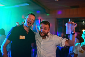 Zwei Männer feiern bei Partylicht; einer hält ein Glas, beide lächeln in die Kamera, im Hintergrund Musiker und Deko.