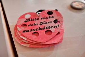 Runde pink bedruckte Karte mit Lochstanzungen und Clip, mit Text „Bitte nicht Herz dein Herz aus­schüt­ten“ als Warnschild.