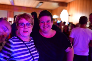 Zwei lächelnde Frauen stehen bei einer Feier im Vordergrund, buntes Licht akzentuiert ihre Gesichter; Hintergrund voller Gäste.