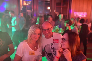Zwei Personen posieren lächelnd bei einer Feier im Vordergrund; im Hintergrund leuchten Scheinwerfer und eine Gruppe im Partyraum.