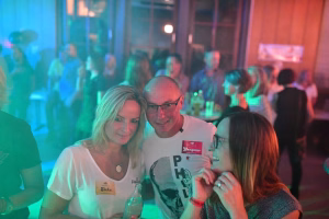 Zwei Personen sitzen eng beieinander bei einer Festveranstaltung; farbiges Bühnenlicht im Hintergrund, Flasche und Pult/Gläser auf dem Tisch.