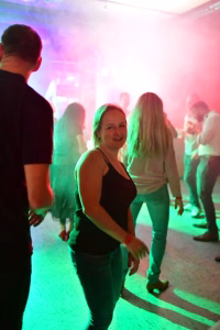 Musikalisch beleuchtete Tanzfläche mit mehreren Personen im Hintergrund; eine Frau dreht sich lachend in die Kamera vor bunten Lichtstrahlen.