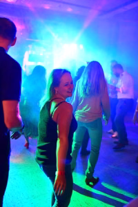 Party-Szene in farbigem Bühnenlicht: zwei Personen tanzen, Blickkontakt, im Hintergrund weitere Gäste und eine Lichtanlage.