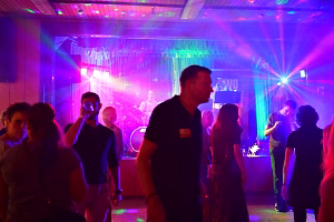Festsaal mit Live-Musik: Gäste vor der Bühne im Vordergrund, farbiges Licht und Nebel, Gitarrist und Schlagzeuger im Hintergrund.