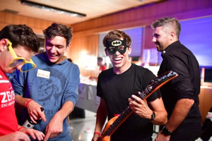 Junge mit Batman-Maske jubelt mit Guitarre in der Hand; weitere Personen schauen und lachen bei einer Party im Raum.