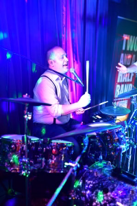 Sänger am Mikrofon mit Drumsticks am Schlagzeug, beleuchtet von blau-roten Bühnenscheinwerfern; wirkt bei Live-Auftritt spielend und singend.