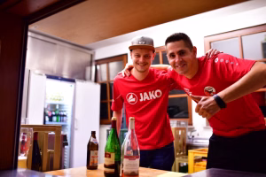 Zwei Personen in roten Shirts mit einem Kappenmann posieren im Innenraum neben Flaschen und Kartons, Finger zeigen zur Kamera.