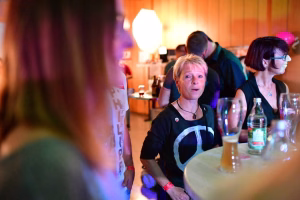 Festsaal mit mehreren Personen; eine Person sitzt am Tisch und blickt zur Seite, im Hintergrund freundliche Gespräche und Deko-Lichtstimmung.