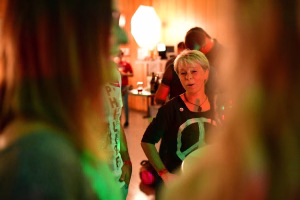 Teilnehmende Person im Innenraum einer Feier, im Schein farbiger Lichter in Gesprächs- oder Ansprachehaltung. Hintergrund mit Tisch und Dekoration.