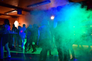 Menschen tanzen in einem Veranstaltungsraum, beleuchtet von farbigen Bühnenlichtern mit starkem Nebel und Sitzbänken im Hintergrund.