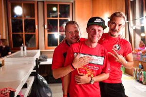 Drei Männer in roten T-Shirts posieren lächelnd und winken in einer Partyhalle; einer hält eine Urkunde, im Hintergrund Tischdekoration.