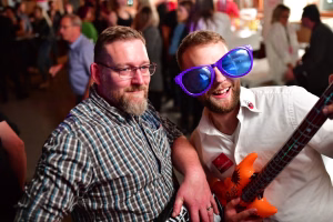 Zwei Männer sitzen im Partyraum, einer mit blauer Partybrille und Lächeln, beide halten eine Requisiten-Geigenmaske und scherzen.