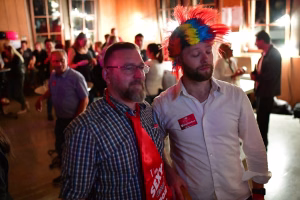 Zwei Männer in festlicher Abendgesellschaft, einer trägt eine bunte Federkopfbedeckung; beide lächeln, während im Hintergrund Gäste sitzen.
