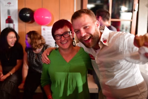 Zwei Personen umarmen sich bei einer Feier; beide lächeln in die Kamera, um den fröhlichen Anlass zu zeigen. Hintergrund mit bunten Luftballons.