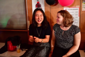 Zwei Frauen sitzen nebeneinander und lachen, eine spricht gestikulierend; Partydekoration und Getränkeliste weisen auf eine Feier hin.
