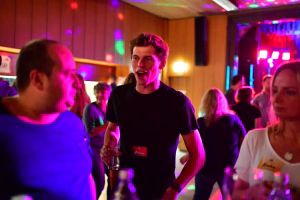 Feiernde Gäste in einer Festhalle, ein Mann in Schwarz lacht und hält ein Getränk, bunte Bühnen-LEDs beleuchten die Tanzfläche.