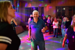 Lächelnder Mann im Partyraum auf der Tanzfläche, umgeben von Gästen; farbiges Bühnenlicht betont die festliche Stimmung.