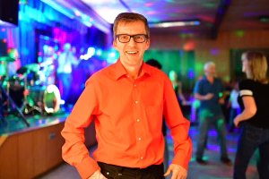 Lächelnder Mann im orangefarbenen Hemd mit Brille vor Bühne, farbigem Licht und Publikum; belebte Atmosphäre einer Feier.