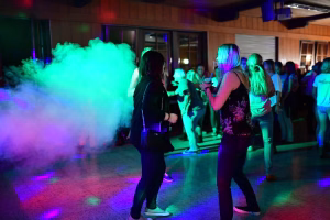 Auf der Partyfläche tanzen zwei Personen im Scheinwerferlicht, während dichter farbiger Nebel die Umgebung einhüllt.