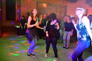 Mehrere Personen tanzen bei einer Feier im beleuchteten Saal, mit farbigen Lichtspots auf dem Boden und Getränken in der Hand.