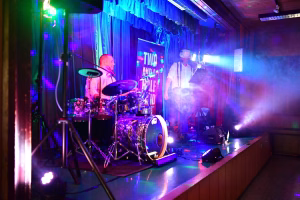 Musikbühne mit Schlagzeuger und Sänger im Scheinwerferlicht; Nebel und blaue Bühnenbeleuchtung unter einem Vorhang für eine Live-Performance.