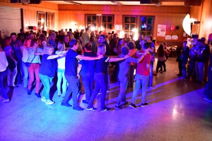 Menschen bilden auf einer Tanzfläche eine enge Reihe, Arme ausgestreckt, vor einer Bühne mit farbigem Licht.