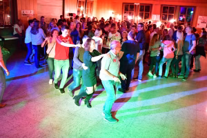 Feiernde Menschen auf einer Tanzfläche bei farbigem Licht, mehrere Paare tanzen vor einer gefüllten Bar.
