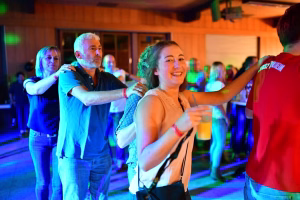 Festsaal bei farbigem Licht: Eine lächelnde Frau mit Handtasche hebt den Arm, im Hintergrund tanzen weitere Gäste.