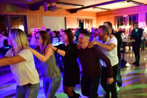 Menschen feiern auf einer Tanzfläche in einem festlich beleuchteten Raum, beugen sich zu einer Gruppe und tanzen im Schein lila Farben.