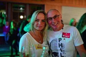 Zwei Personen in weißen T-Shirts, eng beieinander, freundlich in Empfangs-/Feierkulisse mit farbigem Licht dekorativ inszeniert.