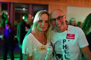 Zwei Personen stehen Schulter an Schulter bei einer Feier, freundlich in die Kamera lächelnd; festliche Beleuchtung im Hintergrund.