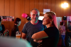 Zwei Personen sitzen beim Fest im Kreis, schauen sich an und lächeln; Tisch mit Getränk, farbiges Licht und Dekoration im Hintergrund.
