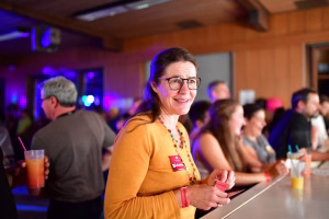 Lächelnde Frau mit Brille an einer Theke bei Veranstaltung; lässiger Plausch, im Hintergrund Gäste im Festsaal bei farbigem Licht.