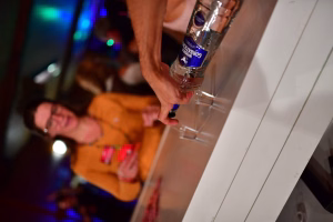 Hand hebt eine Flasche mit blauem Etikett, daneben geht ein anderer Gast unscharf im Festbeleuchtungskontext vorbei.