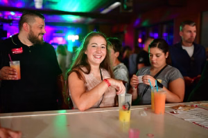 Freundliche Feiergruppe an einer Bar, junge Frau lacht und trinkt, bunte Partylichter im Hintergrund, Getränke und Strohhalme auf dem Tisch.