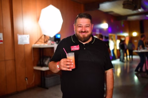 Lächelnder Mann hält ein Glas Drink mit Strohhalm vor festlich beleuchtetem Veranstaltungsraum, im Vordergrund als Gastgebermoment.