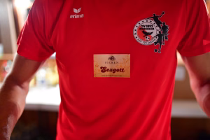 Rotes Vereins-T-Shirt mit Vereinswappen und Aufdruck „125 Jahre“ sowie Anbieter-Label auf der Brust, getragen in unscharfer Umgebung.