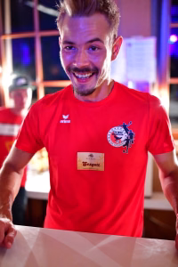 Lächelnder Mann mit rotem Sportshirt steht hinter einem gedeckten Tisch; farbiges Licht im Hintergrund betont die Feststimmung.