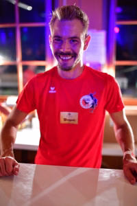 Lächelnder Mann in rotem T‑Shirt sitzt an einem Tisch und hält die Hände an der Tischkante, im Hintergrund bunte Bühnenbeleuchtung.