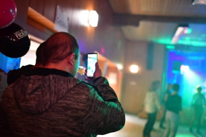 Person fotografiert mit Smartphone im Festsaal; farbiges Bühnenlicht und Deko im Hintergrund, Stimmung und Dokumentation eines Events.