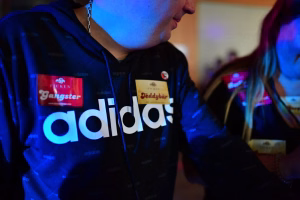 Person trägt eine dunkle Jacke mit großem „adidas“-Logo; im Hintergrund leuchten Banner und Licht, deutet auf ein Event hin.