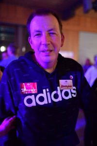 Person in Sportjacke mit violettem Bühnenlicht, hält zwei Aufkleber mit Aufschrift und blickt in die Kamera.