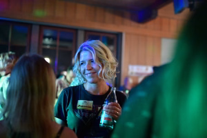 Lächelnde Frau hält ein Flaschenglas bei einer festlichen Feier, während im Hintergrund Menschen und Lichtstimmung zu sehen sind.