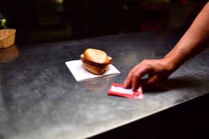 Zwei Würstchen in einem Brötchen auf Papier, daneben legt eine Hand eine rote Kassenscheck-Karte für den Verkauf auf den Tisch.